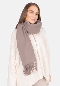Femme portant un pull en maille beige clair et un grand foulard taupe enroulé autour de son cou, avec des franges visibles.
