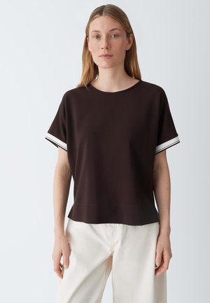 Donna bionda che indossa una maglietta nera a maniche corte con polsini a righe bianche e pantaloni crema a gamba larga, in piedi contro uno sfondo grigio chiaro.