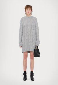 Pulover oversize gri din tricot, cu model de cablu, mâneci lungi și tiv cordonat, asociat cu o geantă neagră și cizme până la gleznă.