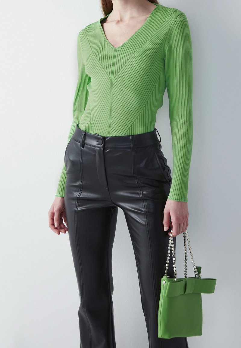 Femme portant un haut côtelé vert vif à col en V, un pantalon en cuir noir taille haute et tenant un sac à main vert assorti avec des anses en chaîne.