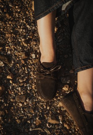 Persona con zapatos mocasines de ante marrón oscuro y vaqueros oscuros, de pie sobre un suelo de grava áspero a la luz natural.