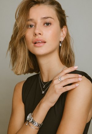 Smykkeegenskaber omfatter en sølvring med sten, lagdelte sølvhalskæder og et sølvur med rem. Modellen har kort, bølget hår.