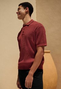 Polo shirt tricotat maroniu, cu o suprafață texturată, mâneci scurte și un guler cu trei butoane, asortat cu pantaloni negri.