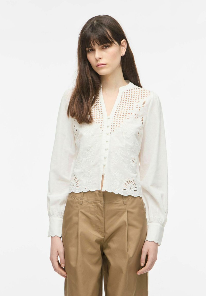 Femme portant un chemisier blanc brodé à manches longues avec des détails découpés et un pantalon plissé beige taille haute, debout devant un fond blanc.