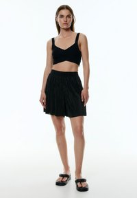 Zwarte geribbelde crop top met brede bandjes, gecombineerd met zwarte geplooide shorts met een elastische tailleband, gedragen met chunky zwarte sandalen.