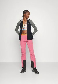 Moteris vilki juodai pilką užtrauktuku užsegamą striukę, gėlių rašto sportinį liemenėlį, rožines kelnes su juodais atvartais ir juodus žygių batus, stovinti su rankomis už nugaros.