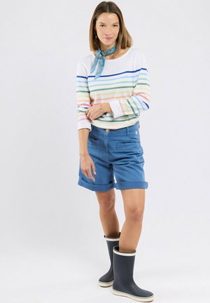 Femme portant une chemise blanche à rayures manches longues, un short bleu, un foulard bleu au cou et des bottes de pluie bleu marine, debout sur un fond uni.