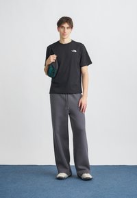 Μαύρο βαμβακερό t-shirt με λογότυπο, γκρι φαρδιά φόρμα, κρατώντας μια σκούρα πράσινη τσάντα. Τα παπούτσια έχουν χοντρό σχεδιασμό. Απλό φόντο.