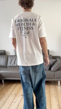 Λευκό μπλουζάκι με μπλε τυπωμένο κείμενο "ORIGINALS HEALTH & FITNESS Club" στην πλάτη. Φορεμένο με φαρδιά τζιν παντελόνια.