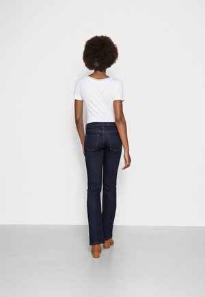 Kvinna med afrofrisyr iklädd en vit kortärmad topp, mörkblå jeans och bruna klackskor, stående vänd mot en vit vägg.