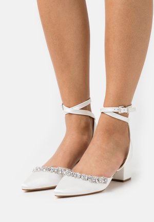 Talons aiguilles en satin blanc avec des brides autour de la cheville et des embellissements en cristal portés sur des pieds nus sur un fond uni.