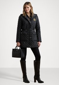 Veste matelassée noire avec capuche, fermetures à boutons-pression et à glissière, emblème doré, deux poches avant. Associée à un jean skinny noir et des bottes montantes.