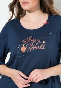 Tummansininen pitkähihaine paita, jossa on painettu kuvio "Ilo maailmalle" punaisella ja kultaisella, korostettuna punaisella nauhalla.