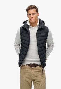 Sort puffervest med vertikalt quiltet mønster, lynlåslommer og hætte, parret med en lysegrå sweater og beige bukser.