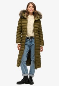 Olijfgroene pufferjas met een met bont gevoerde capuchon, gecombineerd met een crèmekleurige kabelgebreide trui en opgerolde lichtblauwe jeans. Zwarte veterschoenen.
