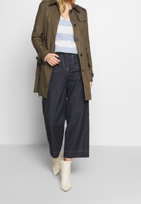 Un trench coat verde oliva sopra un maglione a righe blu chiaro, abbinato a jeans a gamba larga in denim scuro e stivaletti beige.