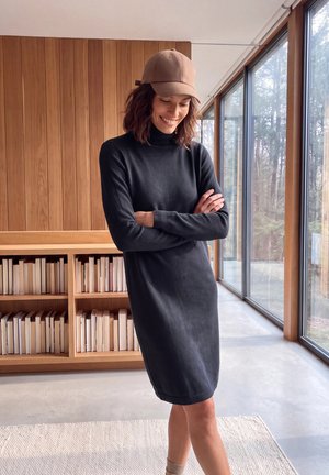 Une femme souriante, vêtue d’une robe noire à col roulé et d’une casquette beige, se tient les bras croisés à côté d’une bibliothèque et de grandes fenêtres.