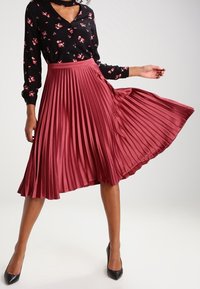 Pleated midi skirt in deep red, made of satin-like material. The fabric has a smooth texture with a fitted waistband and flowing silhouette.