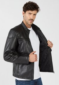 Zwarte leren jas met gestikte schouderaccenten, ritszak op de borst, staande kraag en een gevoerde binnenkant. Draagt open over een wit shirt.