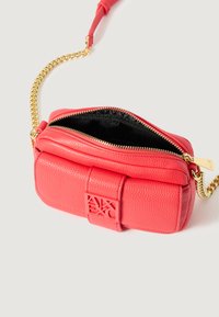 Mała koralowa skórzana torebka crossbody z złotym łańcuszkiem, teksturowana powierzchnia, zamek błyskawiczny oraz wytłoczone logo na przodzie.