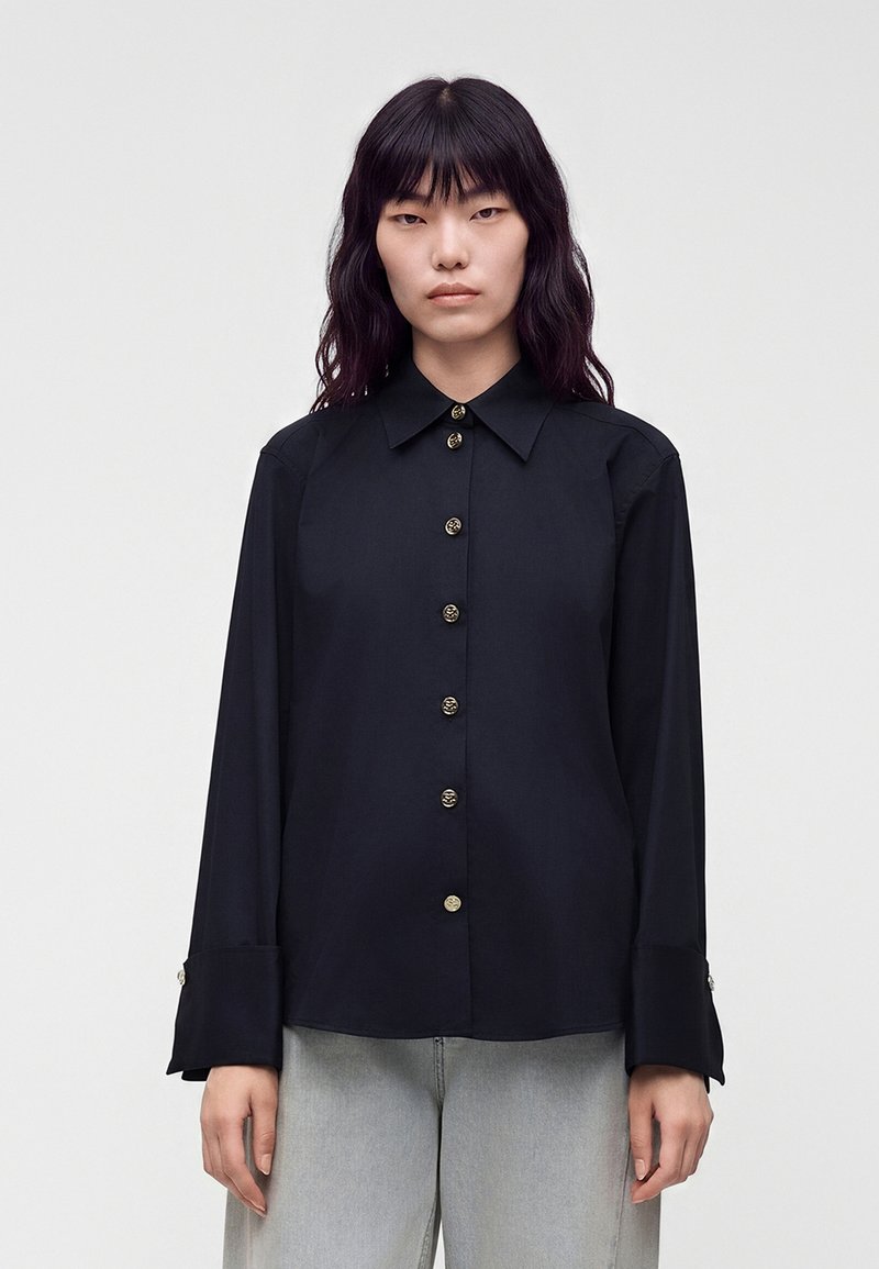 Jeune personne aux cheveux foncés mi-longs porte une chemise noire à boutons et un pantalon gris clair, debout devant un fond clair uni.