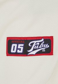 Kreminės spalvos audinys su tekstūruotu raštu. Stačiakampė dėmė rodo "OFFICIAL 05" ir "FUBU" raudona, juoda ir purpurine spalva.