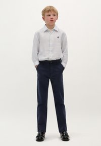 Camisa blanca a rayas con un pequeño emblema, combinada con pantalones navy y zapatos negros, con una textura suave y un corte ajustado.