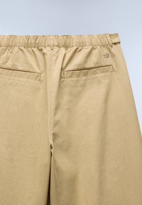 Beige Hosen mit elastischem Bund, zwei paspelierten Gesäßtaschen und einem kleinen gestickten Logo auf der rechten Seite in der Nähe der Taille.