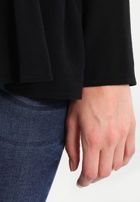 Top in tessuto nero e morbido con una vestibilità ampia, caratterizzato da un design a strati. Mano visibile, indossa jeans di denim blu. Nessun motivo o accessori distintivi.