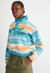 Pullover de fleece em ondas azuis, teal e laranja, com um bolso de peito com zíper. Apresenta um gola alta e botões de contraste.