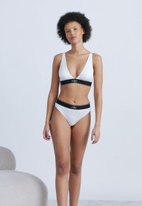 Γυναίκα με σγουρά μαλλιά στέκεται φορώντας λευκό σουτιέν και εσώρουχα με μαύρες ελαστικές ταινίες σε απλό φωτεινό φόντο.