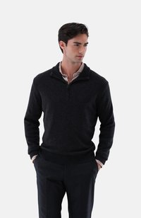 Dunkler Pullover mit Reißverschlusskragen, ribbeltem Saum und strukturiertem Stoff. Getragen über einem gemusterten Hemd, kombiniert mit dunklen Hosen.