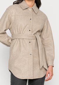 Giacca lunga beige in lana con maniche lunghe e cintura, dotata di colletto e bottoni a pressione frontali, indossata su pantaloni scuri, su un modello con capelli scuri.