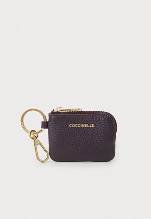 Dark purple leather keychain pouch with a textured finish, gold zipper, and hardware featuring the brand name "Coccinelle" embossed.