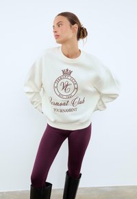 Sweatshirt crema con un logo marrone raffigurante una corona e la scritta "Vermont Club EST. 1991", abbinato a leggings viola scuro e stivali neri fino al ginocchio.