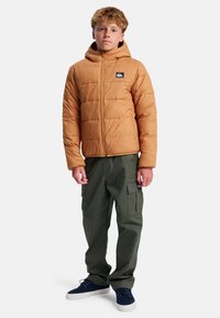 Brązowa pikowana kurtka puchowa z kapturem, zamkiem błyskawicznym i wzorem pikowania. W zestawie z zielonymi spodniami typu cargo i niebieskimi trampkami.