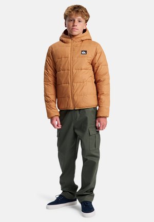 Veste matelassée marron avec capuche, fermeture éclair et motif matelassé. Associée à un pantalon cargo vert et des baskets bleues.