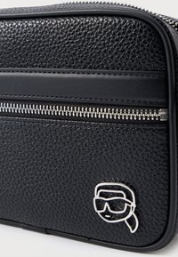 Bolsa sintética texturizada preta com um fecho prateado e detalhe de logo. Apresenta uma faixa de destaque lisa e um pequeno emblema de personagem de desenho animado.