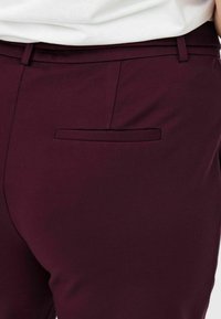 Zbliżenie na bordowe dopasowane spodnie z tylną kieszenią wpuszczaną oraz białą koszulę włożoną w pas.