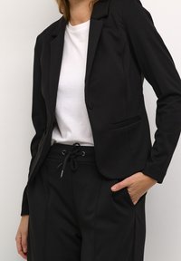 Schwarzer maßgeschneiderter Blazer mit Reverskragen, einer einzelnen Knopfschließe und Fronttasche, kombiniert mit einem weißen T-Shirt und schwarzen Jogginghosen mit Kordelzug.