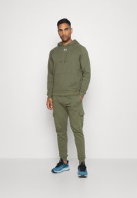 Olivgrüner Hoodie mit einer Fronttasche und Kordelzug, kombiniert mit passenden Jogginghose mit Cargo-Taschen, getragen mit blauen Sportschuhen.