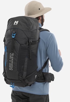 Homme portant un chapeau de soleil beige et une veste bleue, portant un grand sac à dos de randonnée Millet noir avec des accents bleus et de multiples sangles.