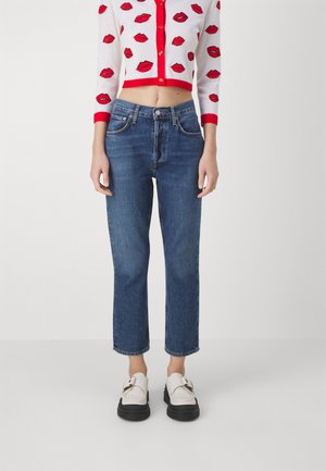 Mujer con cárdigan blanco corto con estampado de labios rojos, jeans azul de talle alto y mocasines blancos con suelas negras, posando sobre un fondo liso.