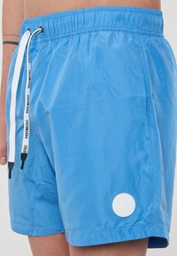 Pantaloni da bagno blu realizzati in materiale leggero e liscio. Presentano una vita elasticizzata con un cordino e un'etichetta circolare bianca sul bordo.