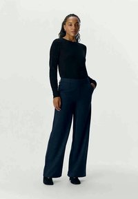 Top de manga longa preto combinado com calças largas azul-escuras, apresentando um tecido suave e detalhes discretos de bolsos. Modelo em pé com as mãos nos bolsos.