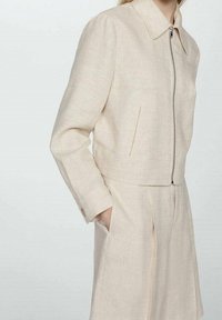 Beige linnedjakke med krave, frontlynlås og sidelommer. Har en cropped design og glat, tekstureret stof.
