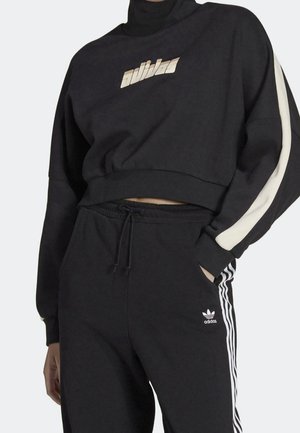 Osoba ubrana w czarny dres Adidas z białymi paskami na spodniach i rękawach, krótka bluza z logo na piersi i kieszenią w spodniach.