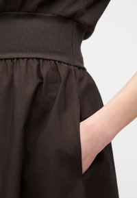 Δεξί χέρι τοποθετημένο μέσα στην πλαϊνή τσέπη σκούρου καφέ φούστας με πιέτες και φαρδιά ριμπ ζώνη μέσης, μπροστά σε απλό φόντο.