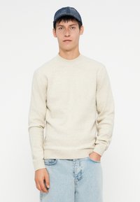 Svetlo bež pleten pulover s okroglim ovratnikom in rebrastimi manšetami, v kombinaciji s svetlo modrimi denim hlačami in strukturirano mornarsko kapo.