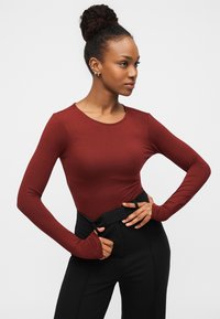 Μακρυμάνικο κορμάκι σε σκουρόχρωμο πορτοκαλί, κατασκευασμένο από ελαστικό ύφασμα, με στρογγυλή λαιμόκοψη και εφαρμοστή σχεδίαση, συνδυασμένο με μαύρο παντελόνι ψηλής μέσης.